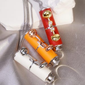 May include: Three pendant necklaces with cylindrical, lipstick-shaped charms in red, orange, and white. Each charm has a decorative top with a dangling design of gold and silver accents, and a silver chain.