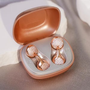 Op de afbeelding: Roze gouden oortelefoonhoesje met twee bijpassende oortelefoons. De oortelefoons hebben een ronde, met kristallen bezette bovenkant en een druppelvormige basis. Het hoesje is open en toont de oortelefoons in een grijze binnenkant.