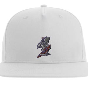 Könnte beinhalten: Weiße Baseballkappe mit flachem Schirm und kleinem gesticktem Design eines Haifischs auf einem Surfbrett. Der Hai ist grau, mit lila und roten Akzenten auf dem Surfbrett und Design.