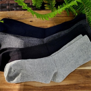 Op de afbeelding: Vier paar geribbelde sokken in verschillende kleuren: marineblauw, grijs, zwart en lichtgrijs. De sokken zijn netjes gerangschikt op een houten oppervlak, met groene bladeren op de achtergrond.