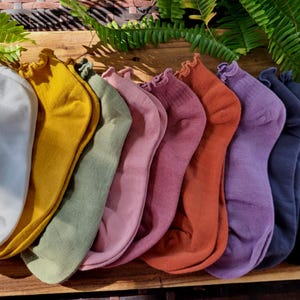 Puede incluir: Una colección de ocho calcetines tobilleros en varios colores, incluyendo blanco, amarillo, verde, rosa, naranja, morado, azul marino y negro. Cada calcetín tiene un borde con volantes y está dispuesto sobre una superficie de madera, con follaje verde en el fondo.