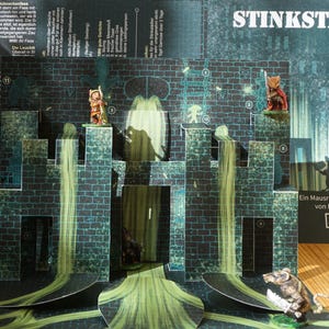 Może przedstawiać: Wyskakująca diorama średniowiecznego miasta, z projektem ceglanej ściany i zielonymi i szarymi akcentami. Diorama zawiera miniaturowe figurki i słowo "STINKSTADT" na górze. Scena rozgrywa się na drewnianej powierzchni.