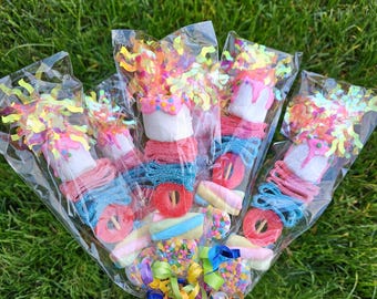 Op de afbeelding: Een boeket van kleurrijke snoepspiesen verpakt in doorzichtig cellofaan. De spiesen bevatten een verscheidenheid aan snoep, waaronder gummy ringen, marshmallows en zure riemen, met kleurrijke confetti en linten.