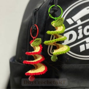 May include: Two spiral-shaped crochet keychains attached to a black backpack. One keychain features red and cream yarn with a red bell, while the other uses green and cream yarn with a green bell. Both have a small leaf detail at the top.