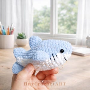 May include: A light blue and white crocheted shark toy with black accents. The shark has a smiling mouth and a black eye. The text "DuyCrochetART" is visible at the bottom of the image.