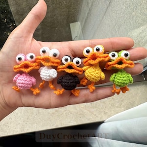 May include: Five small, handmade crochet ducklings in pink, white, black, yellow, and green, each featuring large plastic googly eyes and a bright orange beak. The miniature figures are arranged in a row, showcasing detailed yarn textures.