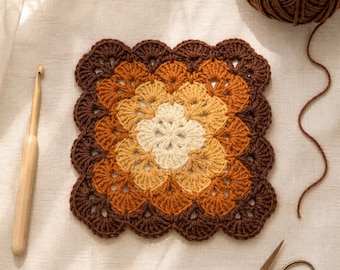 May include: A square crochet motif featuring a scalloped edge in a gradient of cream, golden yellow, burnt orange, and dark brown yarn. A wooden crochet hook lies to the left, while a ball of brown yarn and metal scissors are positioned nearby.