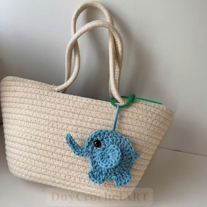 Può includere: Una borsa tote in paglia intrecciata con manici in corda e un ciondolo a forma di elefante blu all'uncinetto. La borsa ha una forma rettangolare e un colore beige chiaro. Il ciondolo a forma di elefante è azzurro con un occhio scuro.