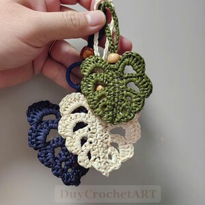 May include: Three crocheted leaf-shaped keychains in navy blue, cream, and olive green. Each leaf has a detailed openwork design. The keychains are attached to a loop with wooden beads and a braided cord. The text "DuyCrochetART" is visible.