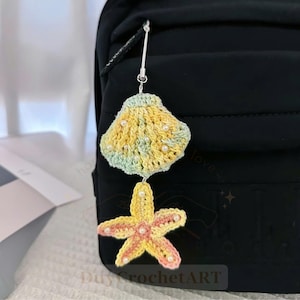 May include: A crochet bag charm featuring a yellow and light green seashell attached to a yellow and pink starfish. Both pieces are embellished with small white beads and connected by a silver-toned metal clasp and chain for attachment.