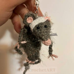 May include: A gray crocheted rat keychain featuring a white bandage wrapped around its head with a small pink spot. The rat has black bead eyes, a pink nose, and pink-tipped paws. It is attached to a silver-toned metal lobster clasp.
