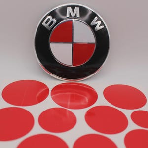 Op de afbeelding: Een BMW-embleem met een zwarte rand, zilveren letters en een rood en wit centrum. Daaronder liggen verschillende rode ovale stickers op een wit vel. Auto-onderdelen.
