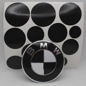 May include: A black and silver BMW emblem with the letters "BMW" and a circular design. A sheet of black circle stickers is behind the emblem. The emblem has a black and silver color scheme.