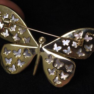 May include: Gold butterfly brooch with a mother of pearl inlay. The butterfly has a detailed design with many small butterflies on each wing.