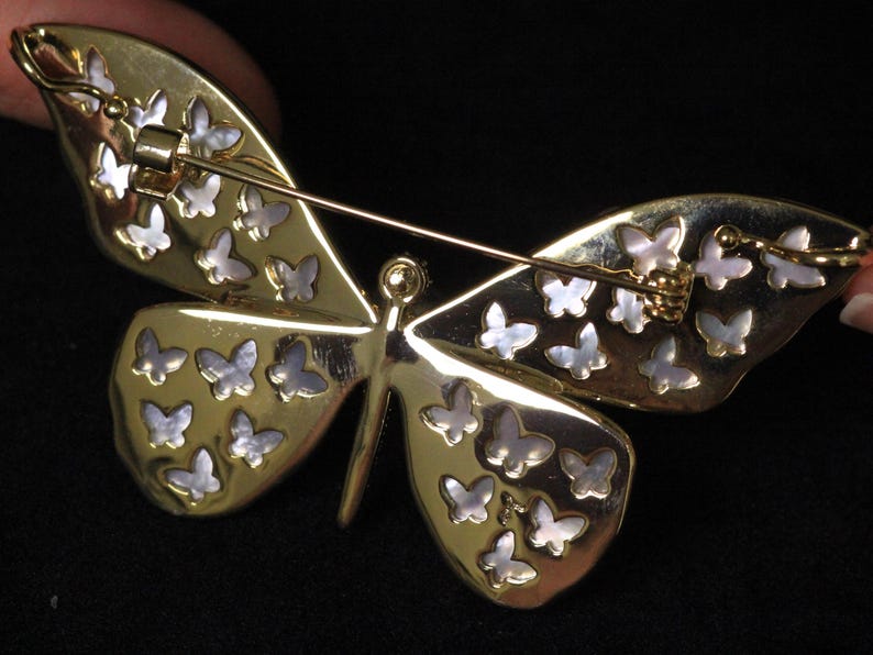 May include: Gold butterfly brooch with a mother of pearl inlay. The butterfly has a detailed design with many small butterflies on each wing.