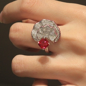 Puede incluir: Un anillo de plata con una gran piedra roja rodeada de piedras blancas más pequeñas. El anillo tiene forma de flor con un borde festoneado.