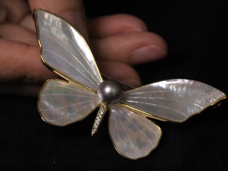 May include: A white mother-of-pearl butterfly brooch with a gold frame and a grey pearl accent.