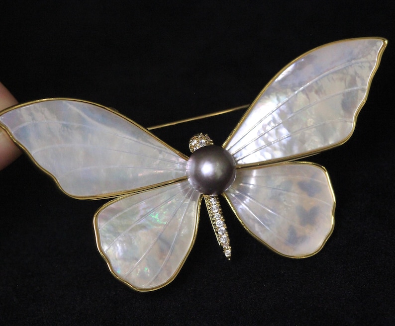 May include: A gold butterfly brooch with iridescent white wings and a dark grey pearl in the centre. The wings are detailed with a delicate gold trim.
