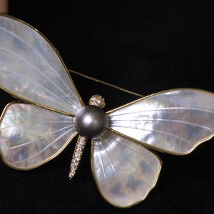 May include: A white mother-of-pearl butterfly brooch with a gold tone finish and a black pearl accent. The butterfly has a detailed wing pattern and a delicate gold tone chain.