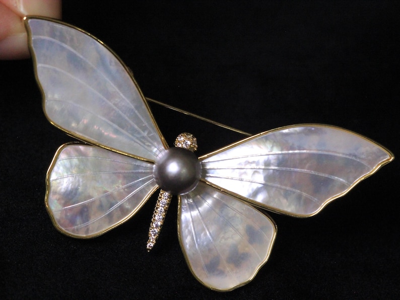 May include: A white mother-of-pearl butterfly brooch with a gold tone finish and a black pearl accent. The butterfly has a detailed wing pattern and a delicate gold tone chain.