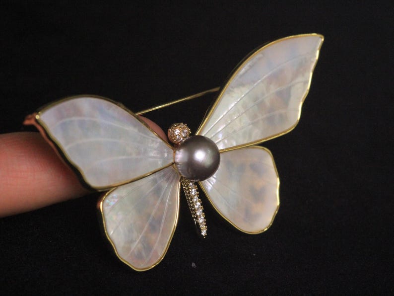 May include: A white mother of pearl butterfly brooch with a dark grey pearl and clear rhinestones.