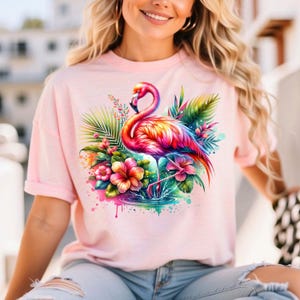 May include: Pink t-shirt with a colorful flamingo and tropical flowers graphic print. The flamingo is standing in a pool of water with splashes of paint.