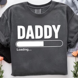 May include: Charcoal grey sweatshirt featuring the word "DADDY" in white, bold capital letters. Beneath the word is a white loading bar with the text "Loading...". The image includes sunglasses, flowers, and a denim shirt.