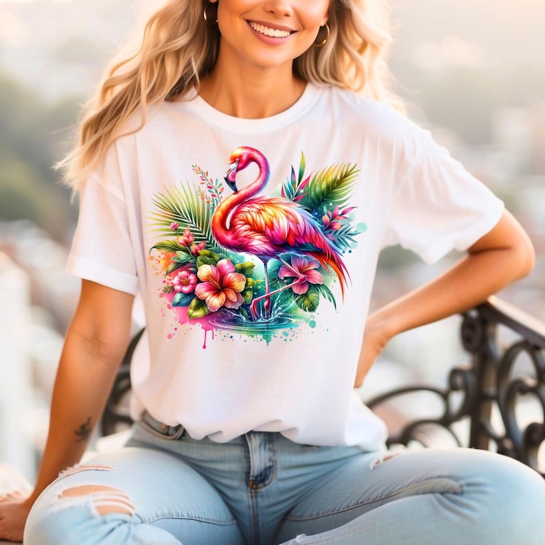 May include: White t-shirt with a colorful flamingo and tropical flowers graphic print. The flamingo is pink, orange, and yellow with a long neck and long legs. The flowers are pink, yellow, and orange. The background is a watercolor wash of pink, blue, and green.