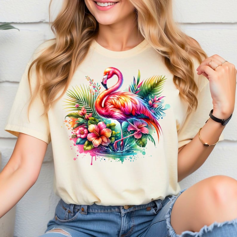 May include: A light yellow t-shirt with a colorful flamingo standing in a pool of water surrounded by tropical flowers and leaves.