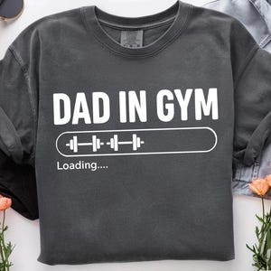 May include: Dark grey sweatshirt with the text "DAD IN GYM" in white, and a loading bar graphic with a barbell. The phrase "Loading...." is below the graphic. The sweatshirt is on a white surface with sunglasses and flowers.