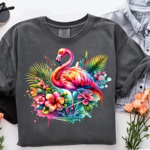 May include: A dark gray t-shirt with a colorful flamingo illustration. The flamingo is standing in a tropical setting with pink, yellow, and green flowers. The design has a watercolor effect.