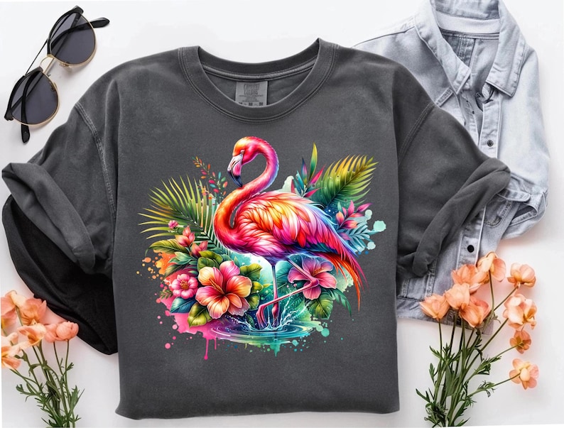 May include: A dark gray t-shirt with a colorful flamingo illustration. The flamingo is standing in a tropical setting with pink, yellow, and green flowers. The design has a watercolor effect.