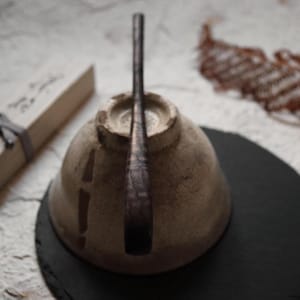 May include: A handmade ceramic teacup with a dark brown handle, resting on a black surface. The cup has a beige, textured finish. A small, rectangular box is visible in the background.