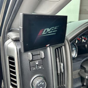 May include: A black Edge monitor installed in a vehicle's dashboard. The screen displays the Edge logo in white and red. The monitor is mounted above the vehicle's control panel, which includes buttons and dials.
