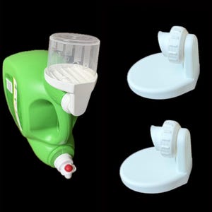 May include: A green plastic bottle with a white dispenser and a red cap. Two white plastic wall mounts are also visible. The dispenser has a clear, graduated cylinder on top. The items are set against a black background.