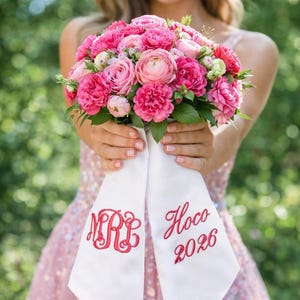 May include: A bouquet of pink roses and other flowers is held in front of a person. The bouquet is tied with a white sash embroidered with the initials "MR" and the words "Hoco 2026" in red.