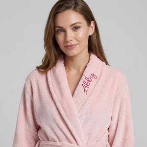 May include: A soft, light pink plush bathrobe featuring a shawl collar and long sleeves. The name Abby is embroidered in a darker pink cursive script on the left side of the collar. The robe has a wrap-front design with a matching fabric tie belt.