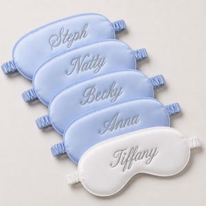 May include: Five satin sleep masks. Four are light blue with names Steph, Natty, Becky, and Anna embroidered in silver. The fifth mask is white with the name Tiffany embroidered in silver. Each mask has an elastic band.