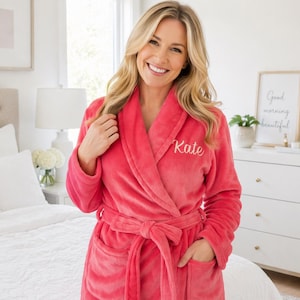 May include: A bright pink plush robe featuring a shawl collar, long sleeves, two front patch pockets, and a matching tie belt. The name Kate is embroidered in white cursive script on the upper left chest area of the soft fabric garment.
