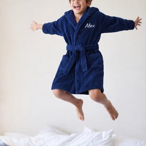 May include: A navy blue hooded bathrobe featuring long sleeves, a tie belt at the waist, and two front pockets. The chest area is embroidered with the name Alex in white cursive script. The robe is made of a soft, textured fabric material.