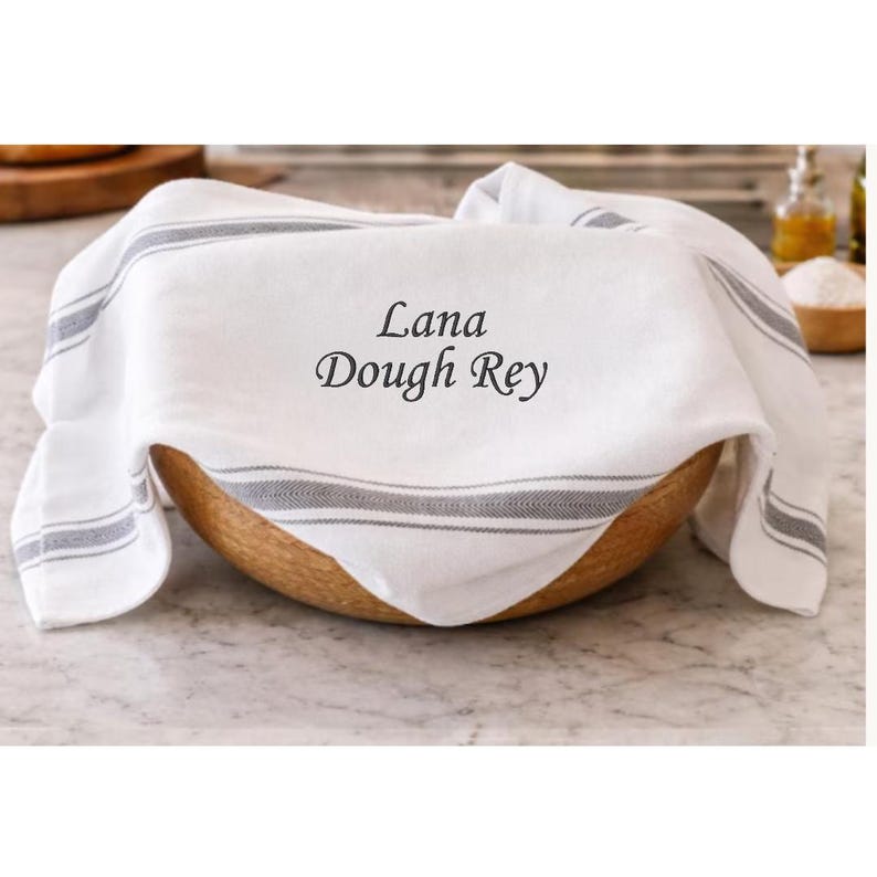 Close-up of a striped bread tea towel embroidered with the starter nickname Lana Dough Rey, covering dough in a wooden bowl on a counter.