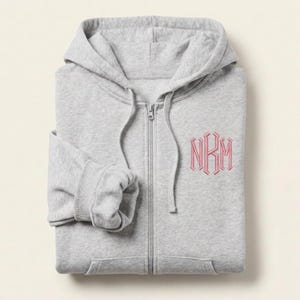 May include: A light gray zip-up hoodie featuring a hood with matching drawstrings and a full-length front zipper. The left chest area displays a pink embroidered monogram with the letters NKM in a decorative, angular serif font style.