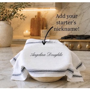 May include: A white kitchen towel with dark gray stripes draped over a round bowl. The towel features the embroidered text 'Angelina Doughlie' in a cursive font. A black arrow points to the text with the caption 'Add your starter's nickname!'