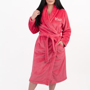 May include: A bright pink plush bathrobe featuring a shawl collar, long sleeves, and a tie belt at the waist. The robe has two front pockets and the name Kate embroidered in white cursive script on the upper left chest area of the garment.