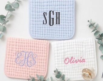May include: Three square, quilted muslin baby burp cloths in light blue, soft pink, and white. The blue cloth features a black embroidered monogram SGH. The pink cloth has a purple embroidered monogram EHS. The white cloth features the name Olivia in pink embroidery and a beige pacifier.