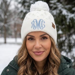 May include: A white knit beanie featuring a cable knit pattern, a large white pom-pom on top, and a ribbed cuff. The front of the cuff displays a light blue embroidered monogram with the letters AMC, and a small brown leather tag labeled C.C.