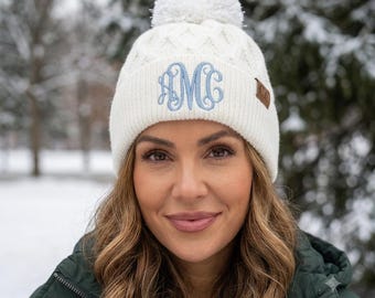 May include: A white knit beanie featuring a cable knit pattern, a large white pom-pom on top, and a ribbed cuff. The front of the cuff displays a light blue embroidered monogram with the letters AMC, and a small brown leather tag labeled C.C.