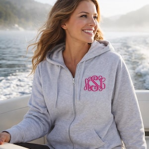 May include: A light gray zip-up hoodie featuring a hood with drawstrings, long sleeves, and two front pockets. The left chest area displays a decorative monogram in bright pink thread, consisting of three intertwined cursive letters.
