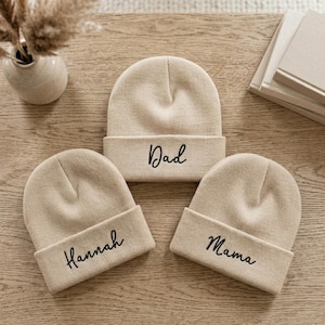 May include: Three beige knit beanies with folded cuffs. Each beanie features black cursive text embroidered on the cuff: one reads Dad, one reads Hannah, and one reads Mama. The beanies are arranged in a triangular formation on a wooden surface.