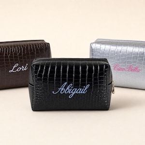 May include: Three rectangular cosmetic bags with a crocodile-embossed texture. The dark brown bag features the name Lori in white, the black bag displays the name Abigail in light blue, and the silver bag shows Ciao Bella in pink text.